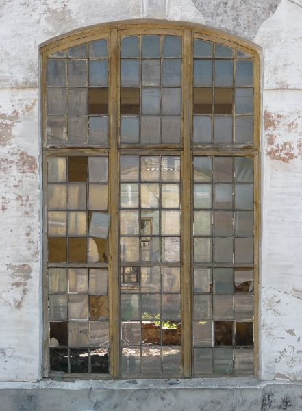 Arched window texture with aged yellow wood separating the small panes, several of which are missing. A whitewashed wall surrounds the window, and a room with debris is visible behind it.