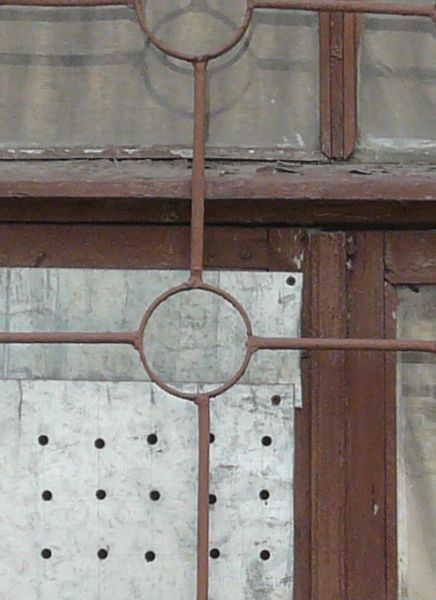 Window texture with red bars formed in a grid and circle pattern over the panes. A white board full of holes covers one pane, and the window is set in a brick wall in various shades of red and yellow.