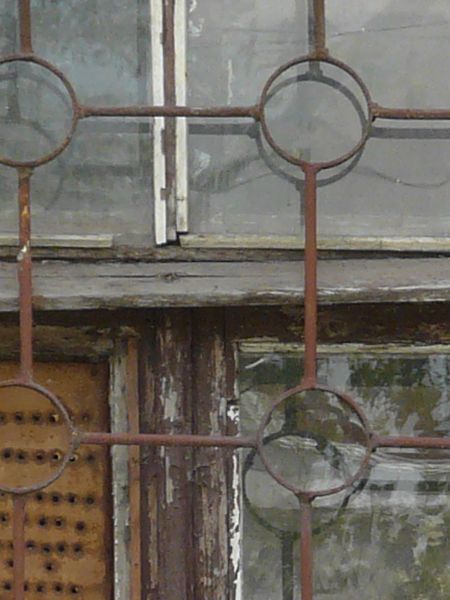 Window texture with red bars formed in a grid and circle pattern over the panes. A white board full of holes covers one pane, and the window is set in a brick wall in various shades of red and yellow.