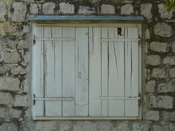 Large white window texture, closed by shutters made up of cracked and broken boards and set in an eroding stone wall. A strip of thin metal runs across the top.