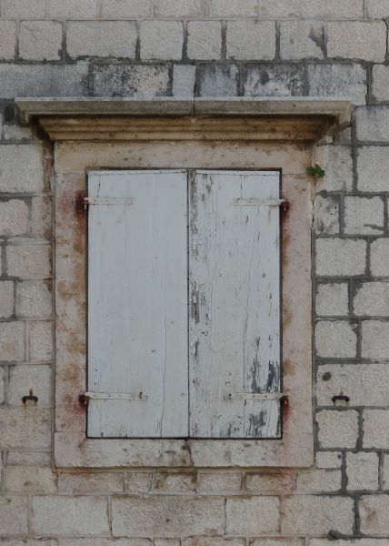 White shutters texture, made of two cracked boards with chipping white paint. A stone frame and brick wall, with brown and black stains, surround the window