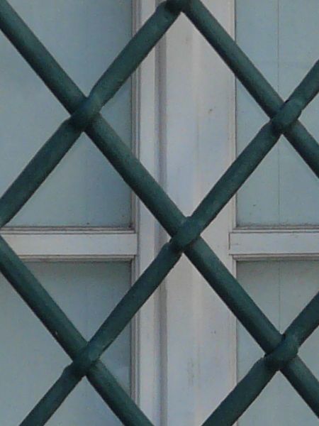 Square window texture, set in a white stone wall, with heavy black bars criss-crossing over it. A white surface is visible behind the window.