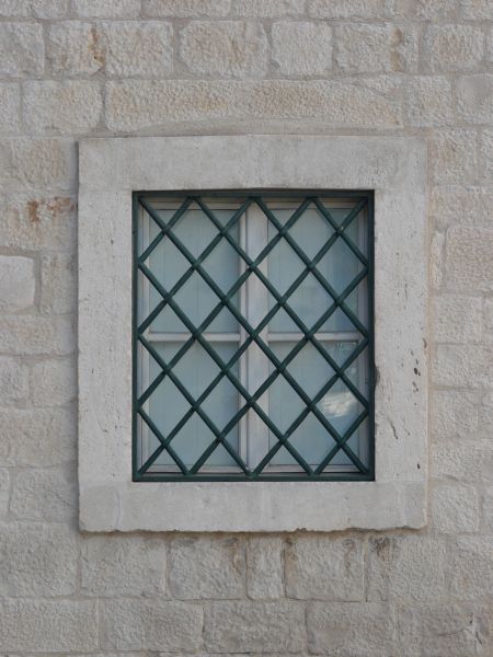 Square window texture, set in a white stone wall, with heavy black bars criss-crossing over it. A white surface is visible behind the window.