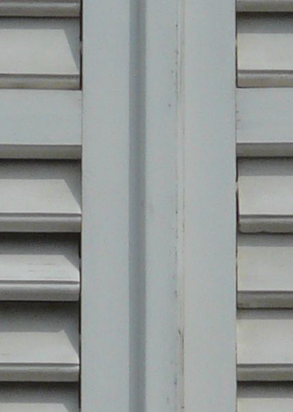 White window texture closed by shutters with brown hinges and very little wear. The window is framed by white stone and set in an eroding grey wall.