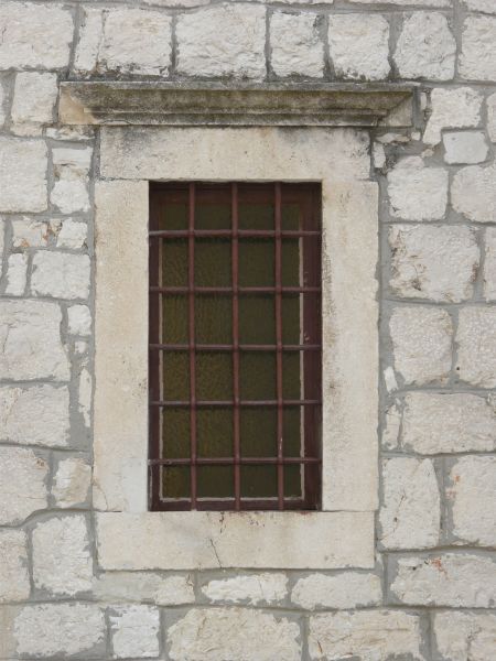 Window texture with red bars set in a grid over the clouded glass. The window is framed by thick white stone with a stained ledge above it, and is set in a white brick wall.