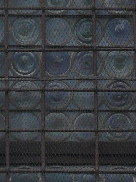 Window texture, with curved stone at the arched top of the frame, set within a light tan brick wall. A uniform pattern of circles is embedded in the glass, on which brown iron bars and wire mesh have been overlaid.