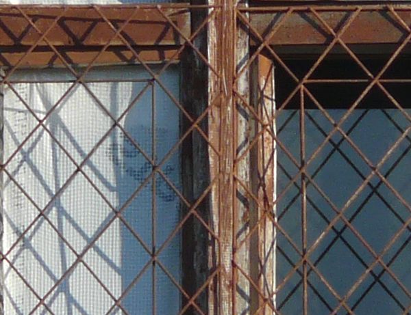 Window texture with panes divided by rusted metal, and partially covered by a rusted metal lattice. White material is visible behind some of the panes, and a white tube is protruding from one of them.