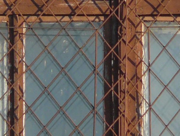 Window texture with panes divided by rusted metal, and partially covered by a rusted metal lattice. White material is visible behind some of the panes, and a white tube is protruding from one of them.