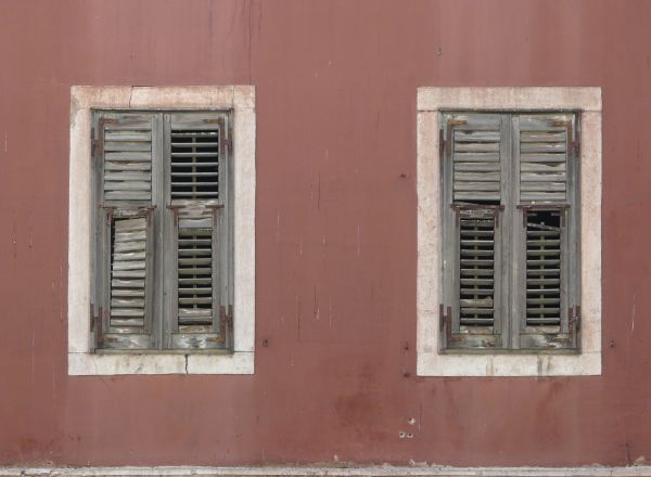 Window texture closed with well-worn grey shutters with rusted hinges. The two windows are surrounded by white stone frames within a slightly stained, rose colored wall.