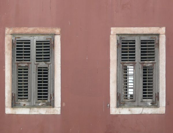 Window texture closed with well-worn grey shutters with rusted hinges. The two windows are surrounded by white stone frames within a slightly stained, rose colored wall.