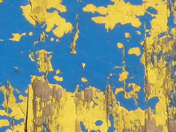 Old wood texture, with chipping layers of blue and yellow paint, and cracked grey boards visible underneath.